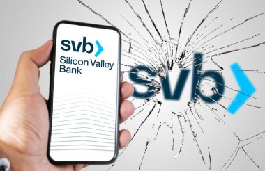 Santa Clara, USA, March 2023: Hand holding a phone with Silicone Valley Bank website on the screen with broken glass and blurred SVB logo in the background. Bank run and bankruptcy. clipart