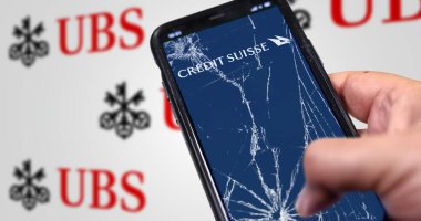 New York, US, March 2023: Hand holding a phone with Credit Suisse logo on cracked screen. UBS logo blurred on white background. UBS bought Credit Suisse for 3B CHF in stock. Illustrative editorial clipart