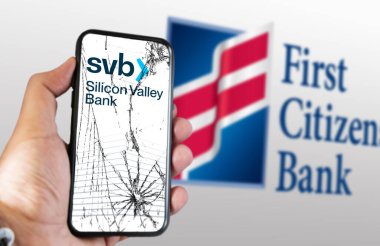 New York, US, March 2023: Hand holding a phone with Silicon Valley Bank logo on cracked screen. First Citizens Bank logo blurred on white background. Illustrative editorial clipart