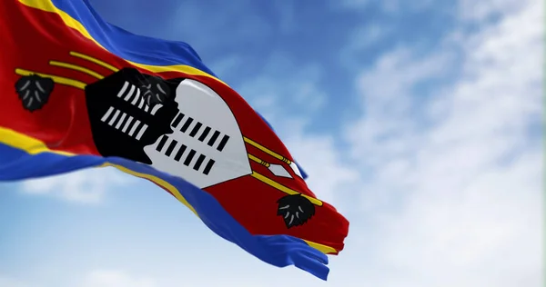 Eswatini national flag waving a clear day. Stripes of blue, yellow, red, yellow, blue with Swazi shield, feathered stick, spears in center. 3d illustration render. Fluttering fabric. Selective focus