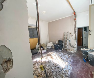 Açık elektrik tesisatı, inşaat aletleri, yıkıntılar ve hasar görmüş duvarlarla dolu bir odanın içinde devam eden yeniden inşa çalışmalarını gösteriyor.