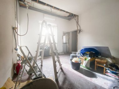 Açık elektrik tesisatı, inşaat aletleri, yıkıntılar ve hasar görmüş duvarlarla dolu bir odanın içinde devam eden yeniden inşa çalışmalarını gösteriyor..