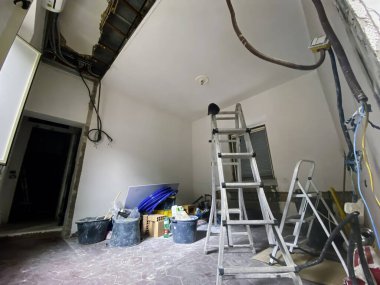 Tadilat sürecinin ortasında bir oda, merdiven, açık elektrik kabloları, inşaat malzemeleri ve yere saçılmış aletler..