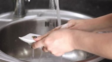 Kadın elleri, akan suyun altında beyaz bir tabağı köpükle yıkıyor.