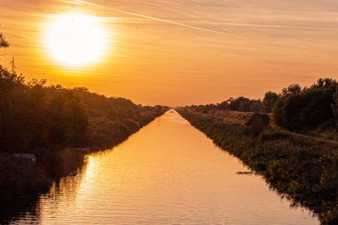 Çarpıcı bir günbatımı, turuncu, pembe ve morun canlı tonlarıyla gökyüzünü boyar. Altın ışık manzaranın üzerine yansıyarak huzur verici ve büyüleyici bir manzara yaratıyor. Mükemmel bir an, günün sonunda güzelliği yakalıyor.