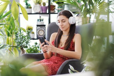 Yeşillik çiçeklerle çevrili evde dinlenen genç kadın kulaklık takıyor ve akıllı telefonuyla iletişim kuruyor.