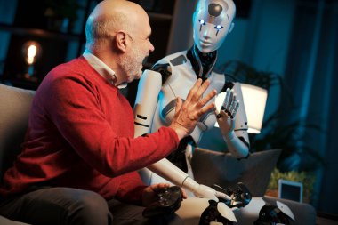 Kıdemli insan ve android robot evde birlikte vakit geçiriyorlar, bir beşlik çakıyorlar.