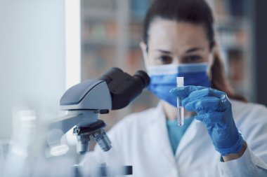 Laboratuvarda çalışan kadın bilim adamı, cerrahi maske takıyor ve mikroskop kullanıyor.