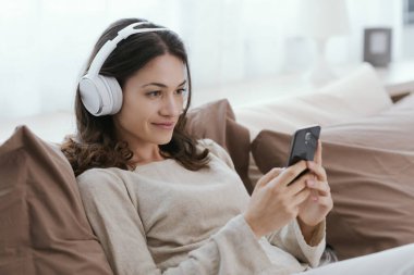 Young woman relaxing on the bed, she is wearing headphones and watching videos on her smartphone