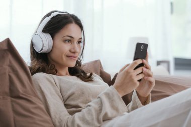 Young woman relaxing on the bed, she is wearing headphones and watching videos on her smartphone