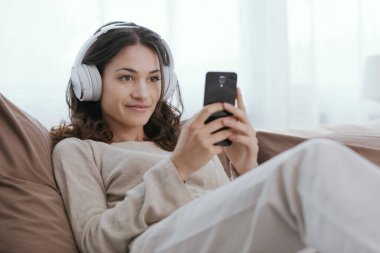 Young woman relaxing on the bed, she is wearing headphones and watching videos on her smartphone