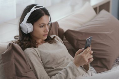 Young woman relaxing on the bed, she is wearing headphones and watching videos on her smartphone