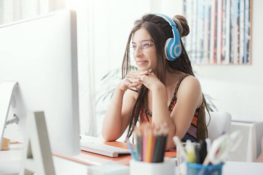 Evdeki masada oturan genç kadın, bilgisayar ekranını izliyor ve kulaklık takıyor.