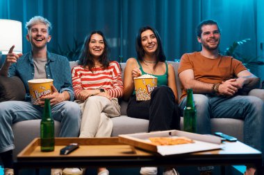 Evdeki kanepede oturan mutlu gençler birlikte yemek yiyor ve film izliyorlar.