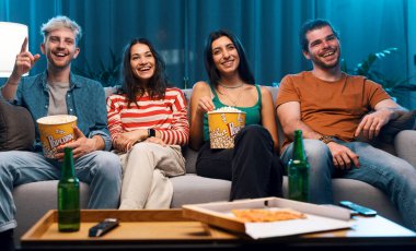 Evdeki kanepede oturan mutlu gençler birlikte yemek yiyor ve film izliyorlar.