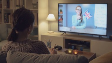 Kanepede oturup televizyon izleyen genç bir kadın, televizyon programlarıyla İngilizce öğreniyor.