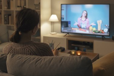 Kanepede oturan ve televizyonda yemek programı izleyen bir kadın yeni tarifler öğreniyor.