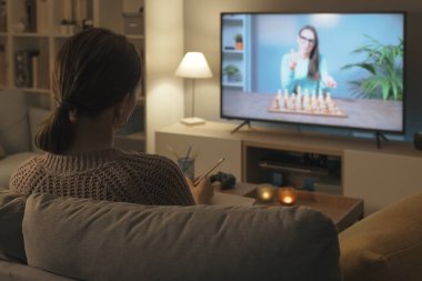 Kanepede oturan ve satranç oynamayı öğrenen bir kadın bir televizyon programı izliyor.