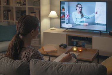 Kanepede oturup televizyon izleyen genç bir kadın, televizyon programlarıyla İngilizce öğreniyor.