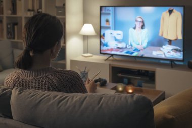 Evdeki kanepede oturan ve televizyonda dikiş kursunu izleyen bir kadın.
