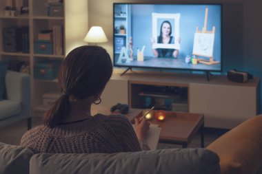 Kanepede oturan bir kadın televizyonunda resim ve sanat kursunu izliyor.