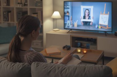 Kanepede oturan bir kadın televizyonunda resim ve sanat kursunu izliyor.