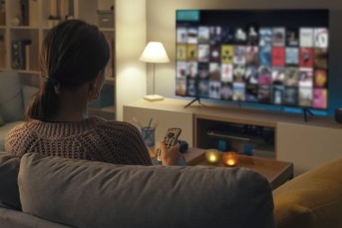 Kanepede dinlenen kadın uzaktan kumandayı kullanıyor ve televizyon menüsünden bir televizyon programı ya da film seçiyor.