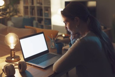 Genç bir kadın masada oturuyor ve evdeki bilgisayarıyla, teknoloji ve iletişim konseptiyle bağlantı kuruyor.