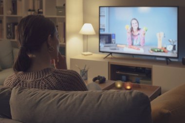 Kanepede oturan ve televizyonda yemek programı izleyen bir kadın yeni tarifler öğreniyor.