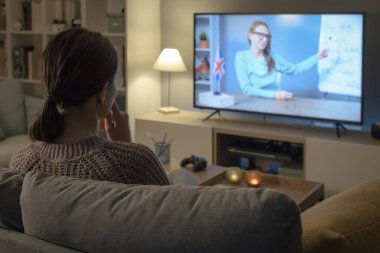 Kanepede oturup televizyon izleyen genç bir kadın, televizyon programlarıyla İngilizce öğreniyor.