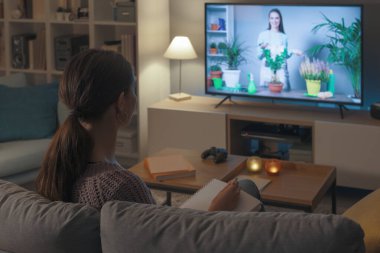Kadın evdeki kanepede dinleniyor ve akıllı televizyonunda bahçe şovu seyrediyor.