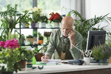 Çiçekçisinde çalışan bir işyeri sahibi faturaları kontrol ediyor ve bir telefon görüşmesi yapıyor.