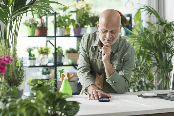 Professional florist working in a plant shop, he is taking orders on his smartphone