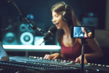 Genç sanatçı stüdyoda şarkı kaydediyor ve akıllı telefonuyla video çekiyor.