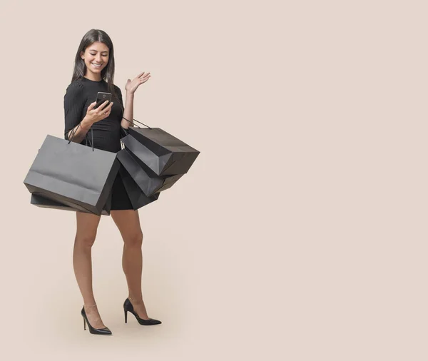 美しいエレガントな女性は ショッピングバッグを保持し 彼女のスマートフォンでオンラインショッピングを行っています ブラックフライデーセール — ストック写真