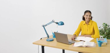 Kendine güvenen kız öğrenci kitap okuyor ve laptopuyla bağlantı kuruyor. Çevrimiçi dersler ve e-öğrenim kavramını takip ediyor.