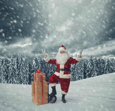 Cesur Noel Baba, karın altında Noel hediyeleri dağıtıyor. Bir el arabasının yanında poz veriyor.