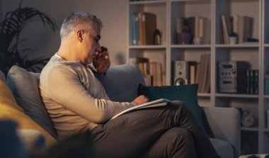 Olgun bir adam evdeki koltukta dinleniyor ve dergi okuyor.