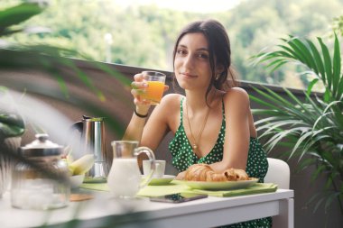 Genç mutlu kadın balkonda oturup kahvaltı yapıyor, portakal suyu içiyor ve kameraya gülümsüyor.