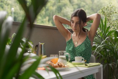 Genç bir kadın balkonda oturuyor ve kahvaltı yapıyor, saçlarını tutuyor.