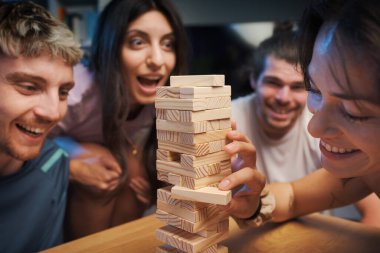 Genç mutlu insanlar evde Jenga oynuyor, bir kız kuleden bir tahta bloğu kaldırıyor ve diğerleri izliyor.