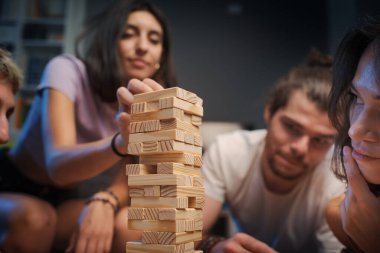 Genç mutlu insanlar evde Jenga oynuyor, bir kız kuleden bir tahta bloğu kaldırıyor ve diğerleri izliyor.