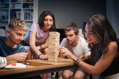 Genç mutlu insanlar evde Jenga oynuyor, bir kız kuleden bir tahta bloğu kaldırıyor ve diğerleri izliyor.