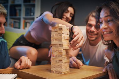 Genç mutlu insanlar evde Jenga oynuyor, bir kız kuleden bir tahta bloğu kaldırıyor ve diğerleri izliyor.