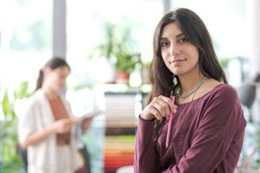 Ofiste poz veren yaratıcı genç tasarımcı kadın kameraya gülümsüyor ve elinde bir kalem tutuyor.