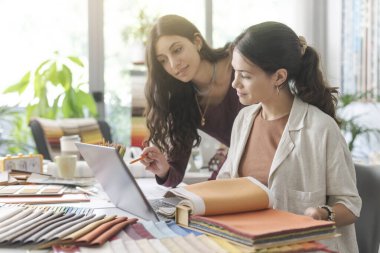 Ofiste birlikte çalışan profesyonel genç iç mimar ve tasarımcılar bir dizüstü bilgisayar kullanıyorlar ve kumaş örneklerini kontrol ediyorlar.