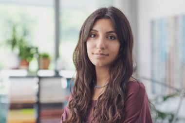 Gülümseyen ve kameraya bakan mutlu genç bir kadın. Arka planda bitkiler olan bir oda.