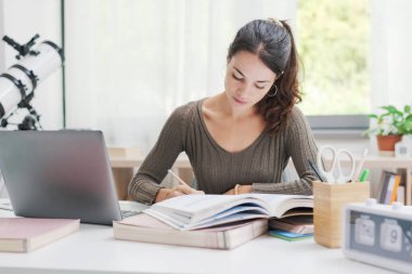 Genç bir kadın evinde oturuyor ve ders çalışıyor. Yazıyor ve dizüstü bilgisayar kullanıyor.