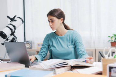 Evdeki masada oturan ve bilgisayarına bağlanan genç bir öğrenci, çevrimiçi dersler alıyor.