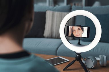 Evde sosyal medya için video çeken genç bir kadın akıllı telefonunu ve halka ışığını kullanıyor.